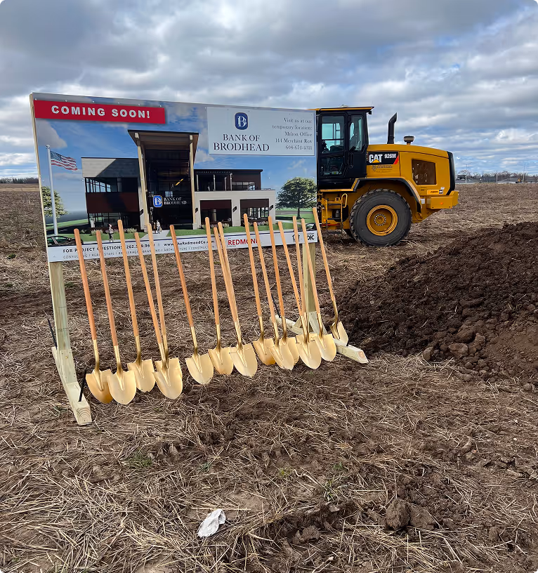 Row of golden shovels lined up in front of a Bank of Brodhead construction sign and a yellow CAT 926M wheel loader at the breaking ground site.