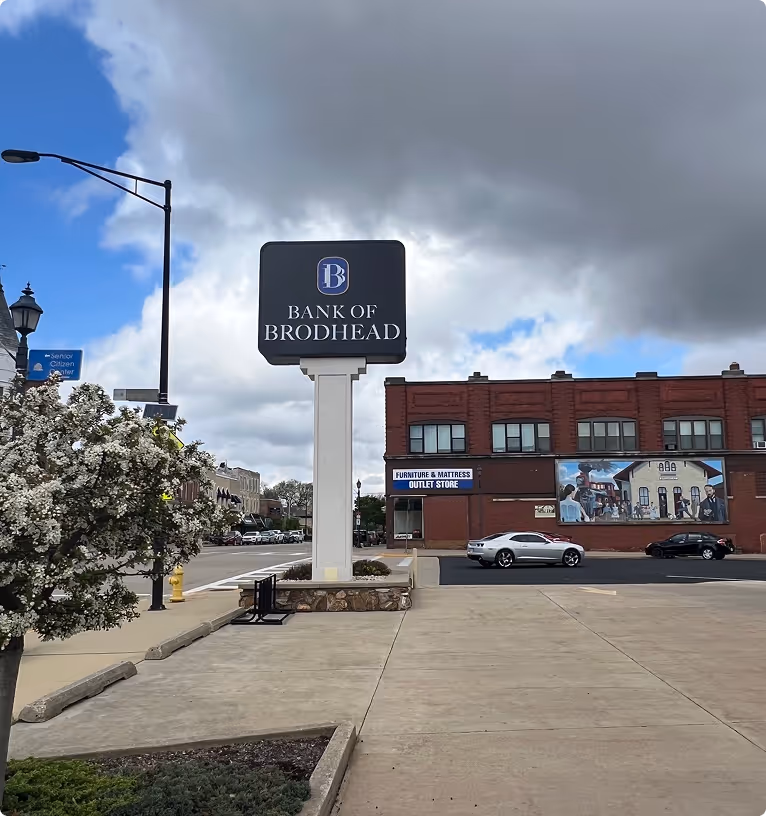 Street view of Bank of Brodhead sign with a cloudy sky, flowering tree, parked cars, and a brick building with a mural.