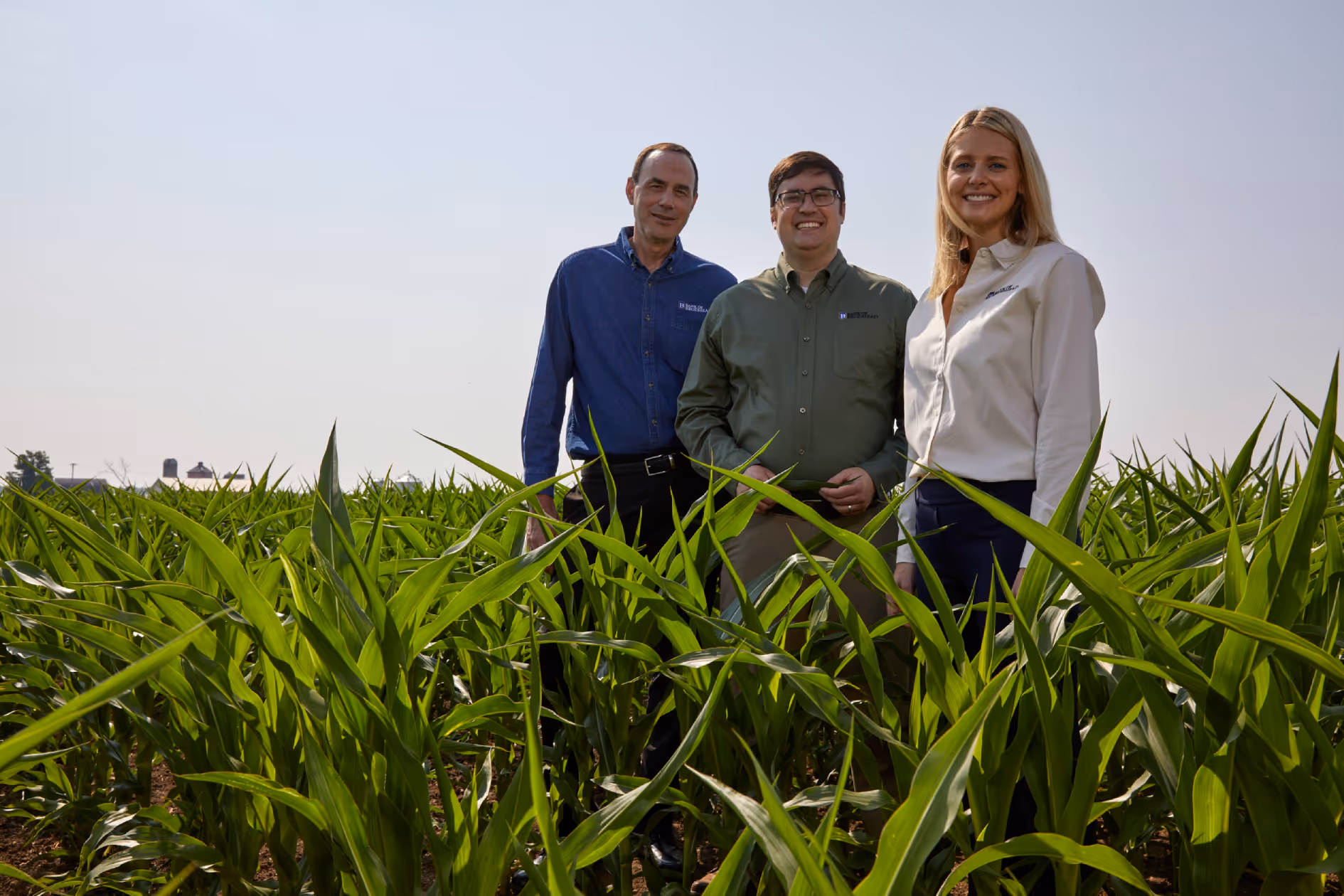 Three people standing in a green cornfield under a clear sky, smiling at the camera.