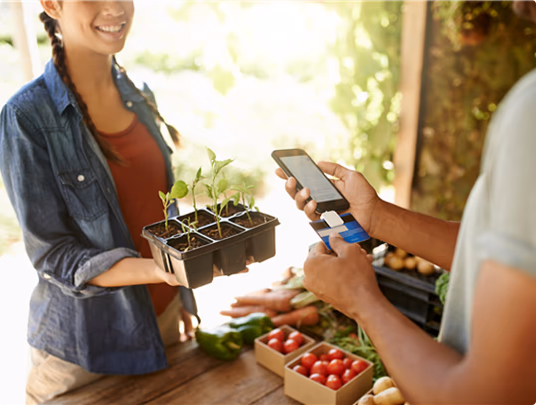 Person with braided hair holding a tray of seedlings while another person makes a payment with a smartphone and card at a market stand with fresh produce.