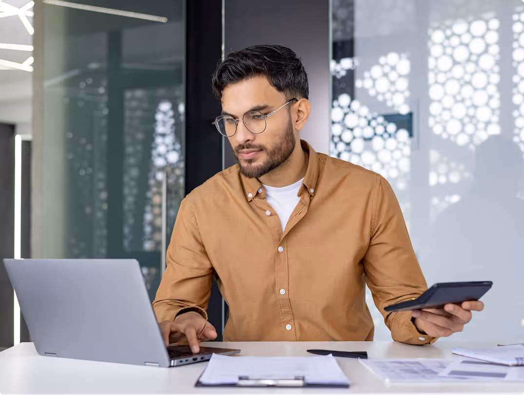 Man wearing glasses working on a laptop while holding a calculator in a modern office.