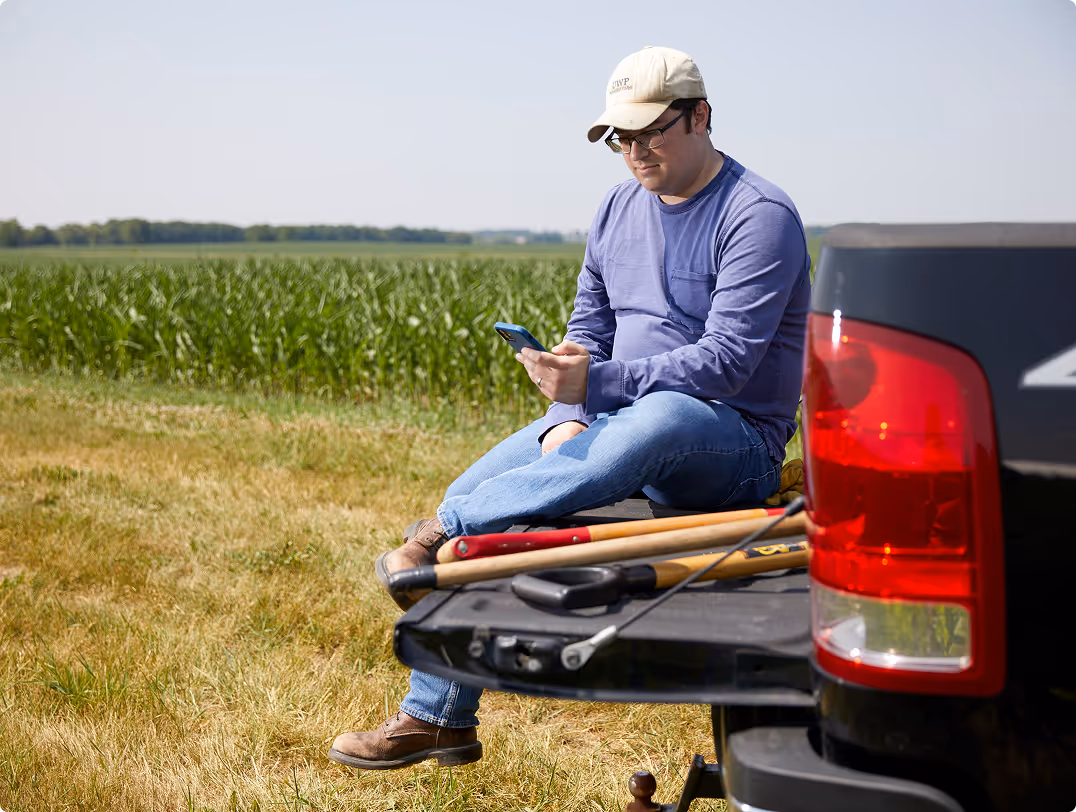 Man wearing a cap and glasses sitting on the tailgate of a pickup truck in a field, looking at his phone with gardening tools beside him.