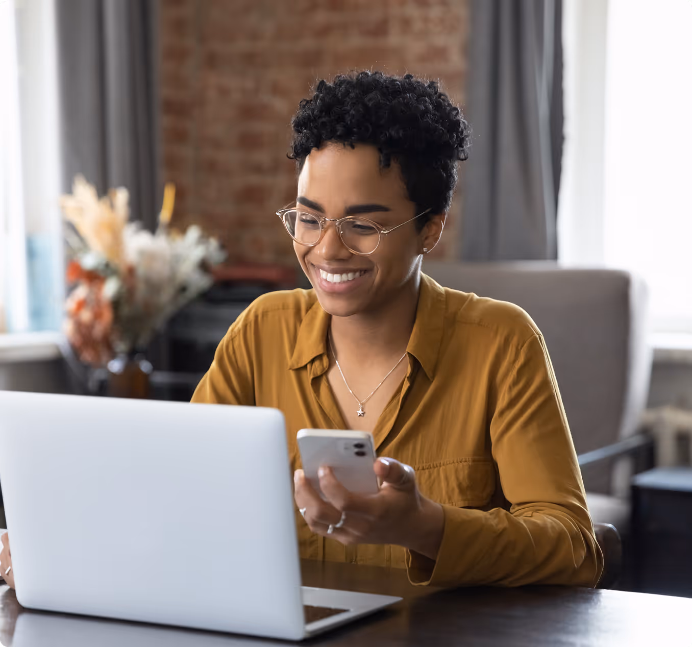 Smiling woman wearing glasses using smartphone and laptop at a table in a cozy room.