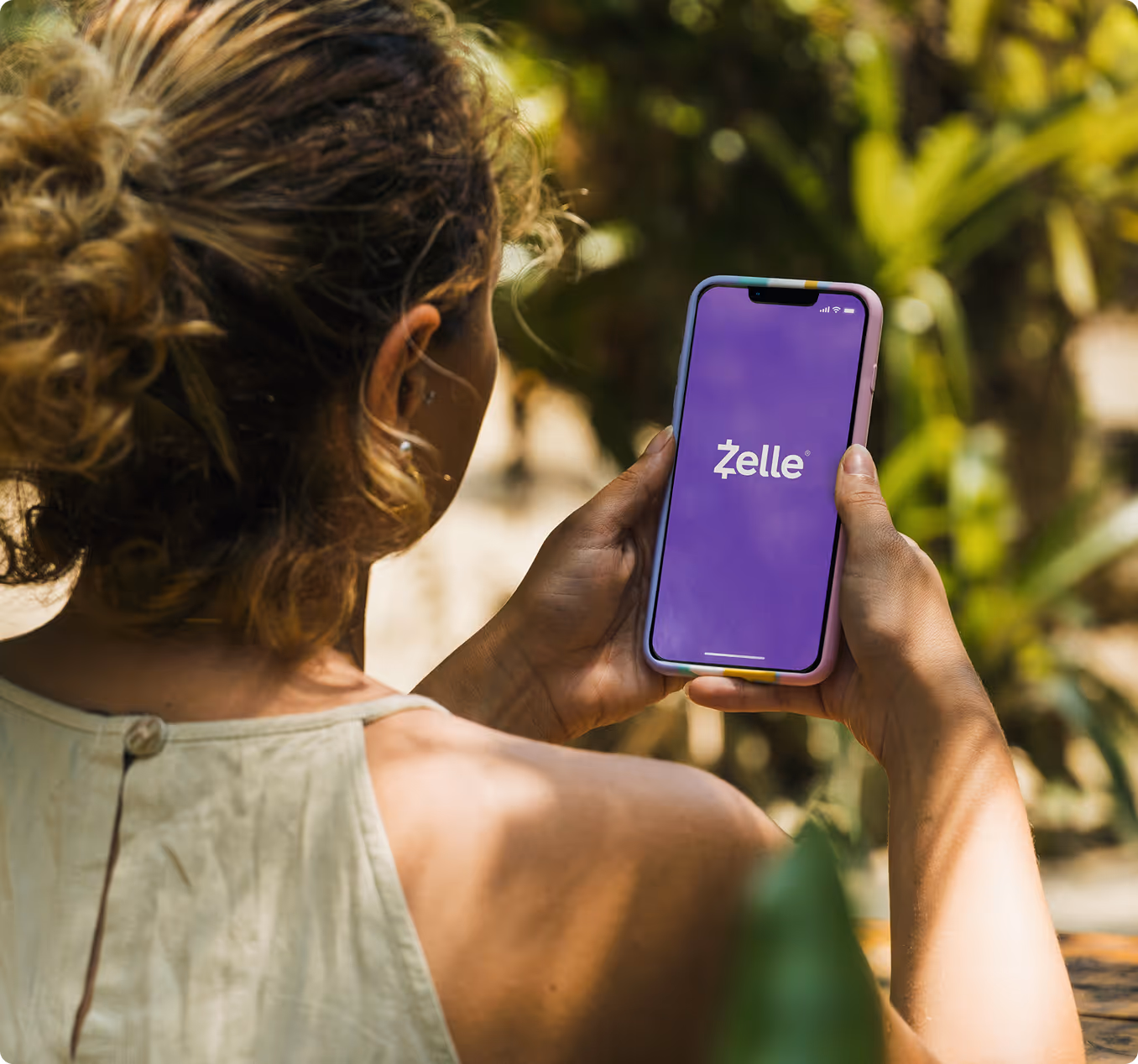 Person holding a smartphone displaying the Zelle app logo on a purple screen outdoors.