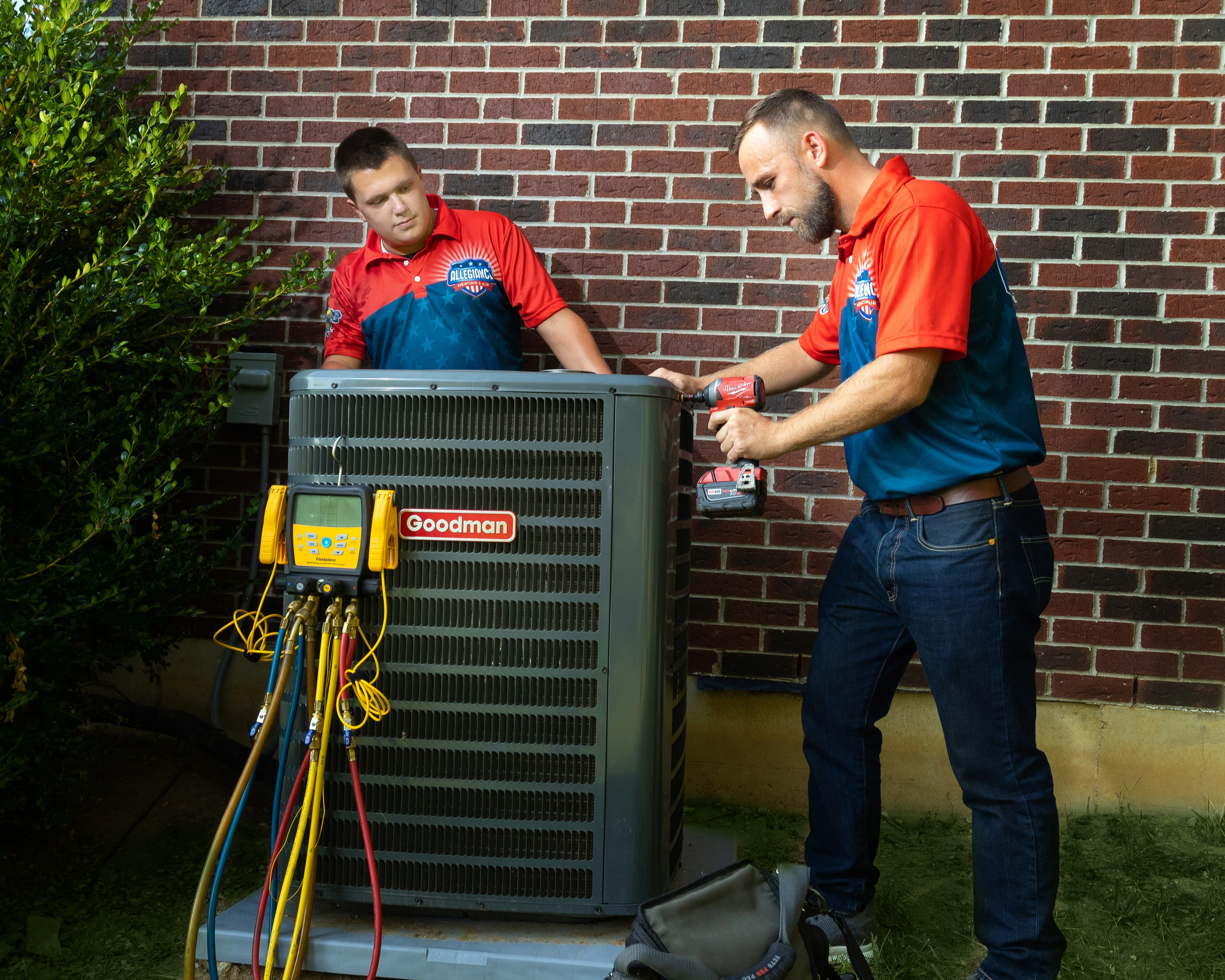 Two technicians in branded shirts repair a Goodman air conditioning unit outside a brick building. 