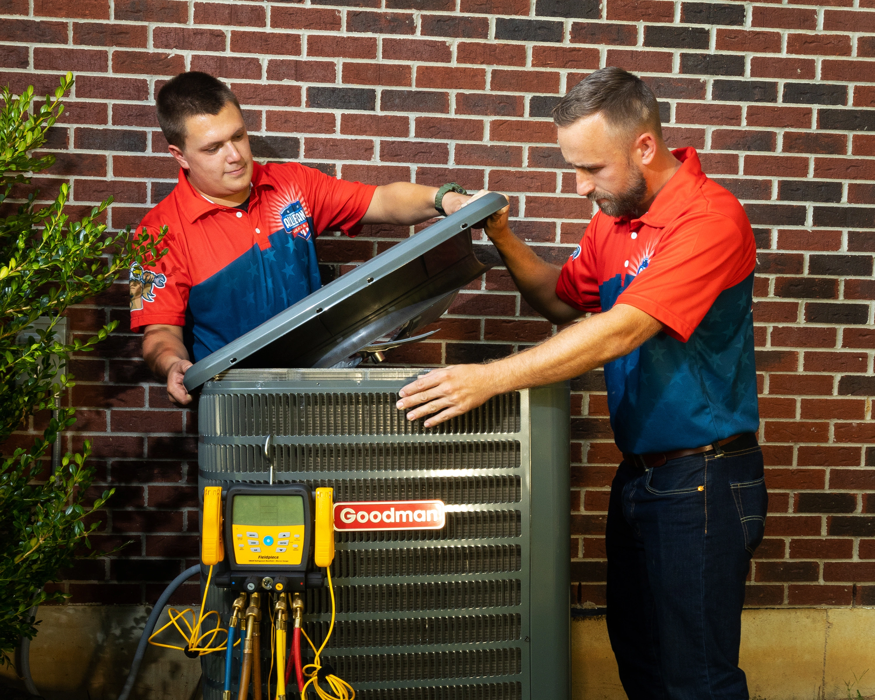 Two technicians in red and blue uniforms work on an outdoor Goodman HVAC unit against a brick wall
