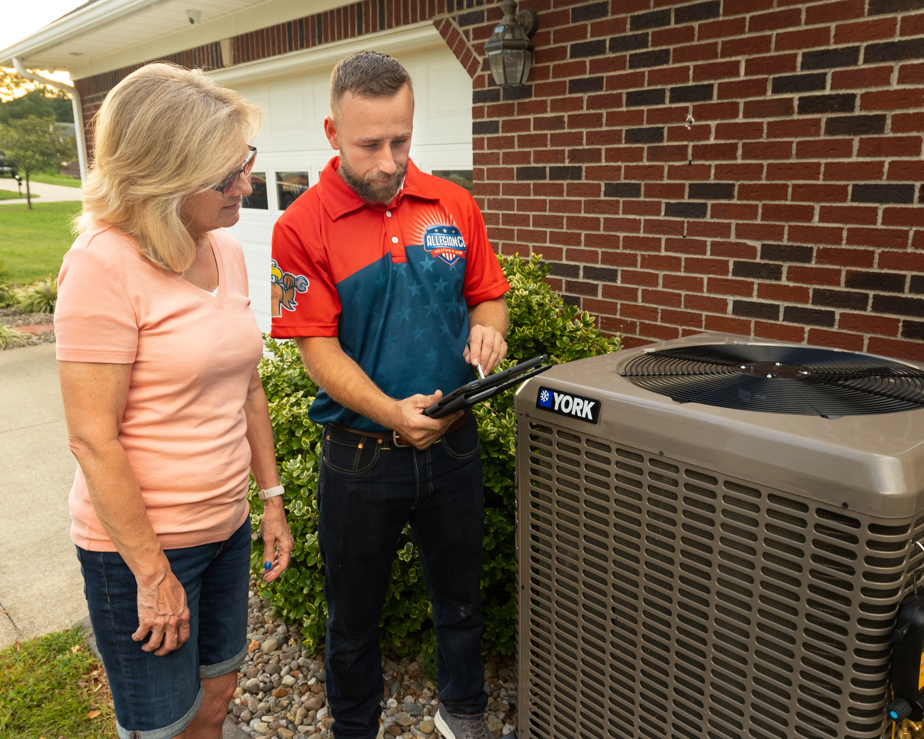 A technician in a red and blue uniform shows a woman something on a tablet next to a York air conditioning unit. 
