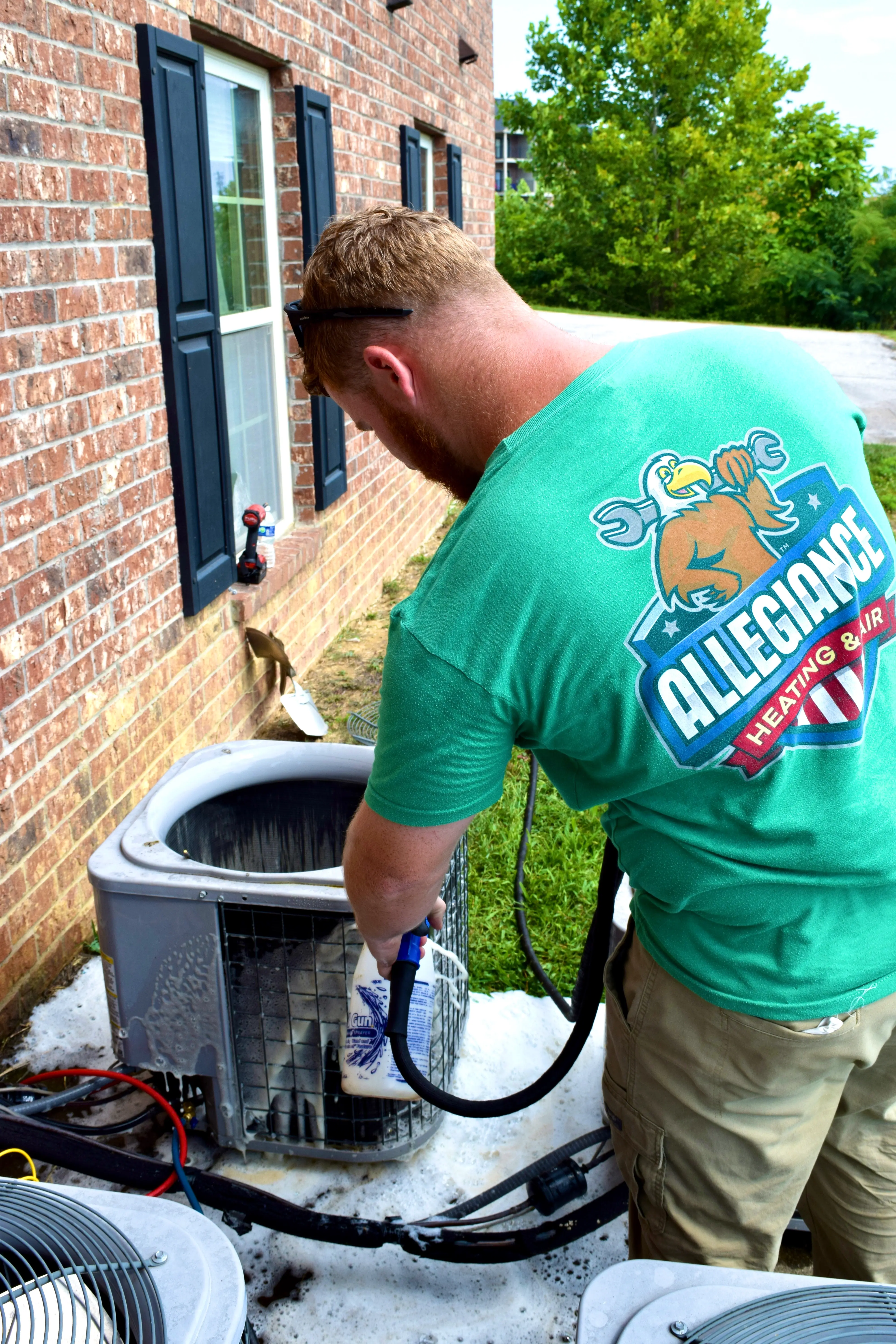 Man in a green "Allegiance Heating & Air" shirt cleans an outdoor AC unit beside a brick house.