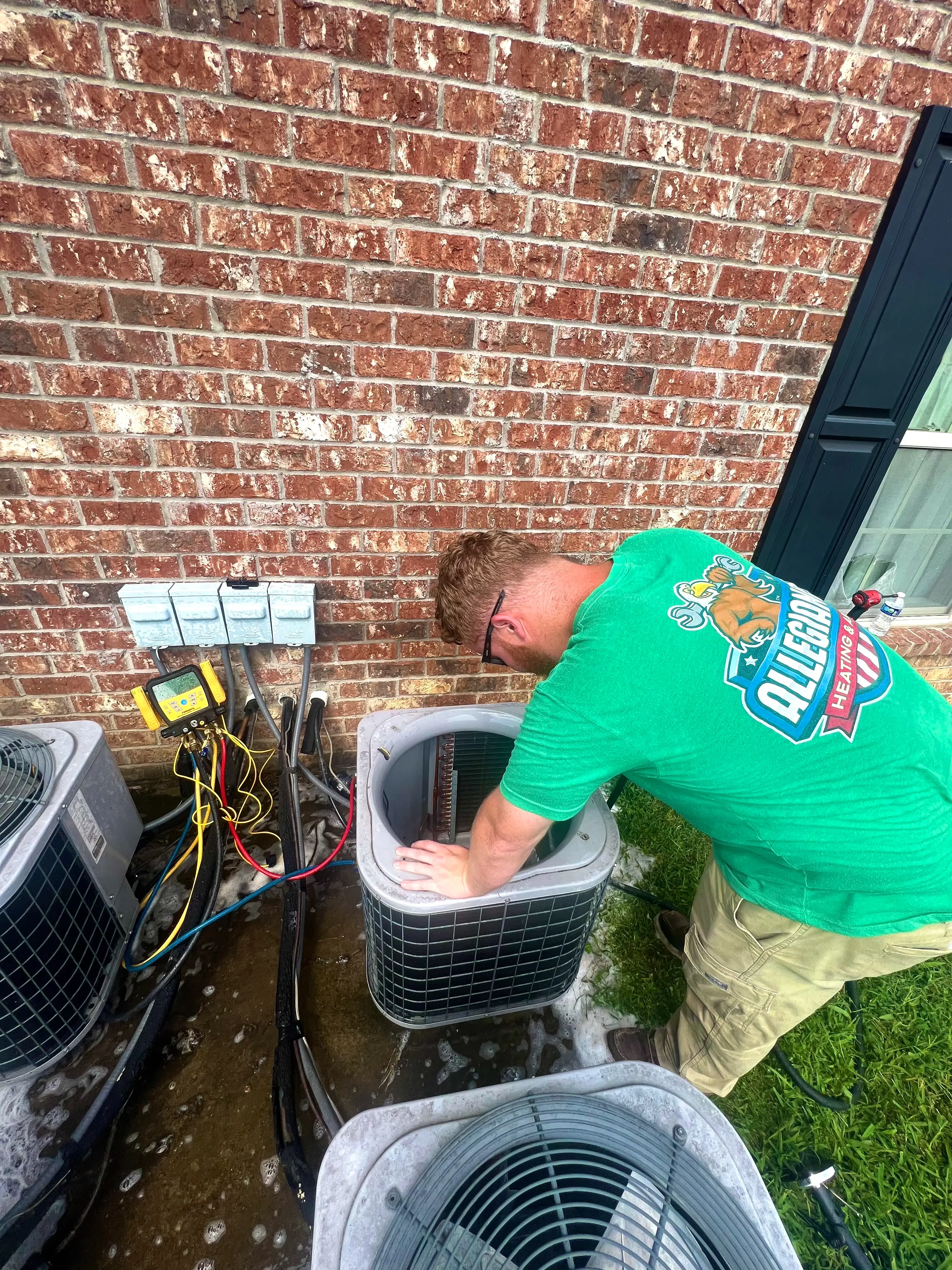 A technician in a green T-shirt works on an outdoor air conditioning unit next to a red brick wall. 