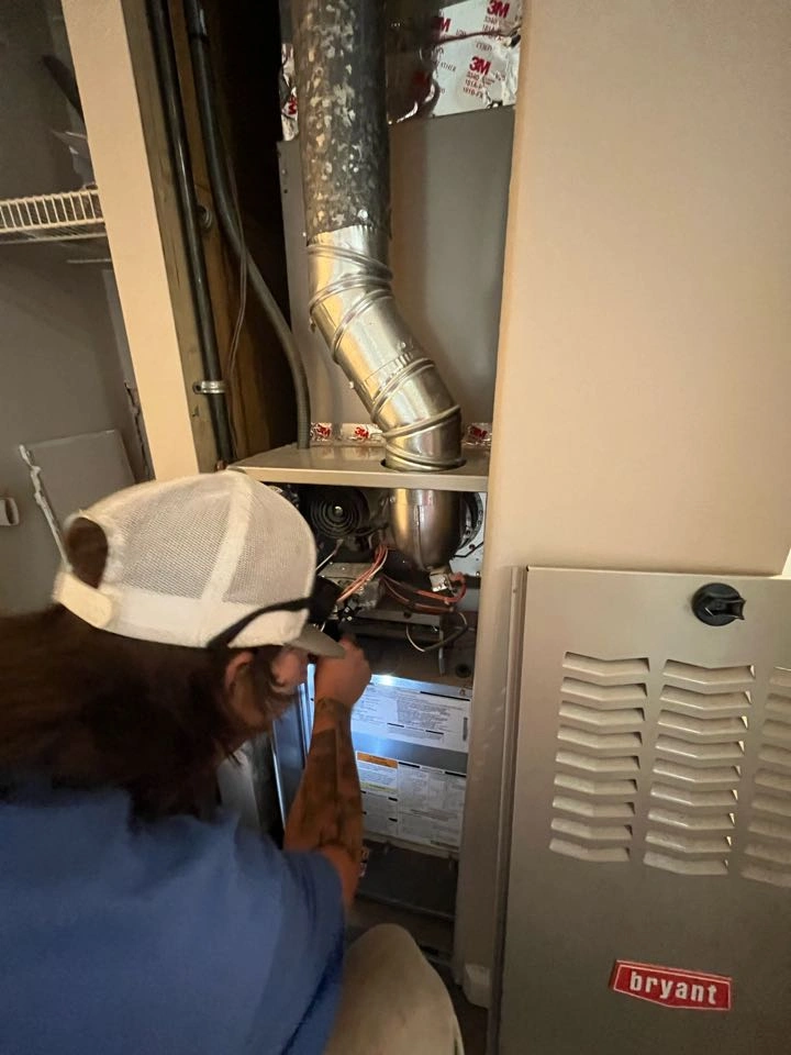 A person in a blue shirt and cap repairs a furnace in a utility closet. 