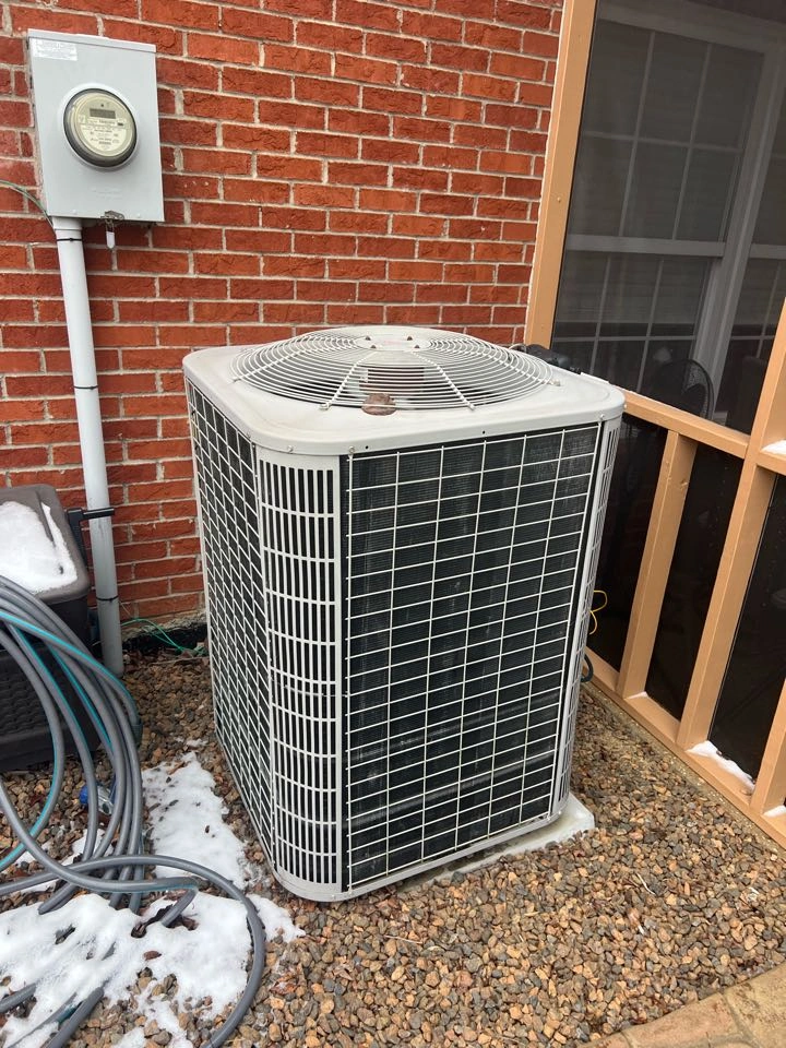 Outdoor air conditioning unit next to a brick wall, surrounded by gravel and some snow.