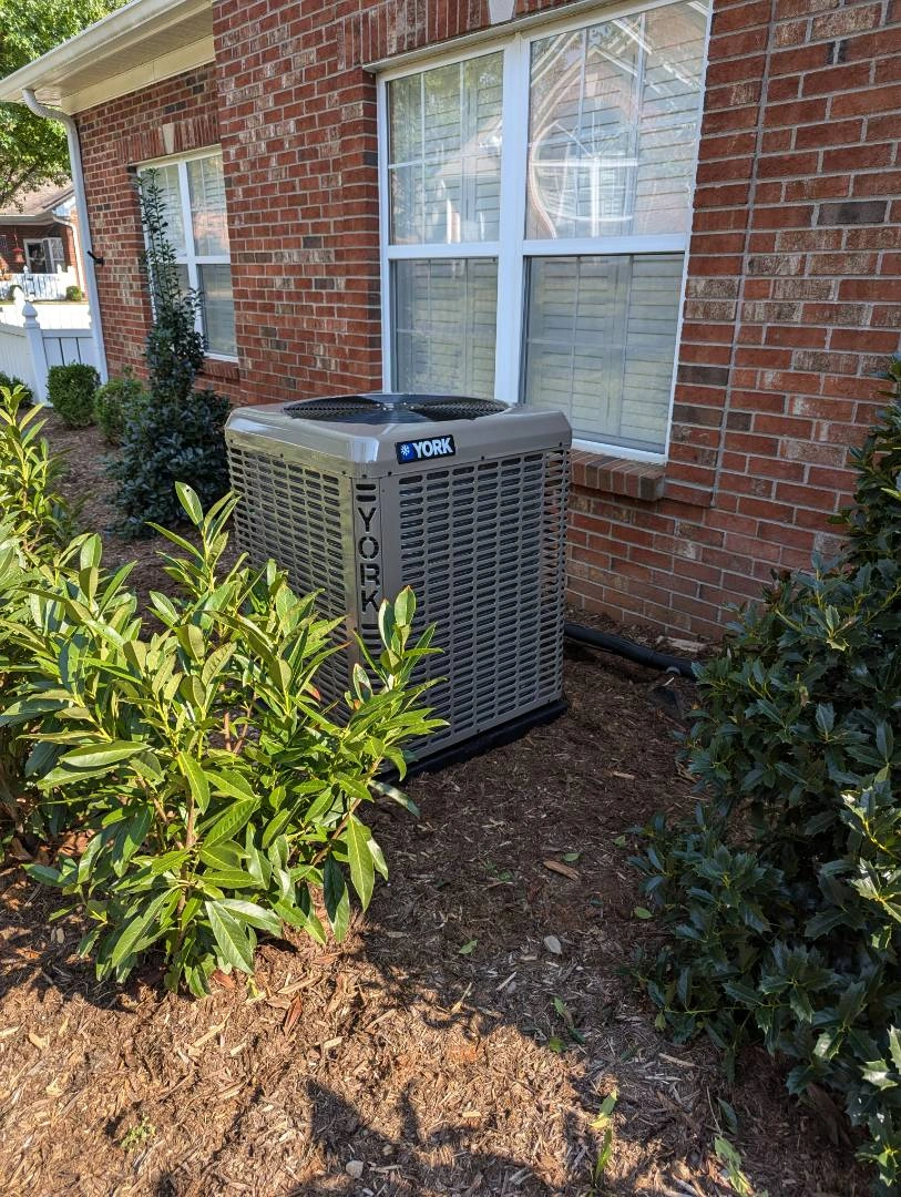 A York air conditioning unit sits on a mulch bed beside a brick house, surrounded by green shrubs.