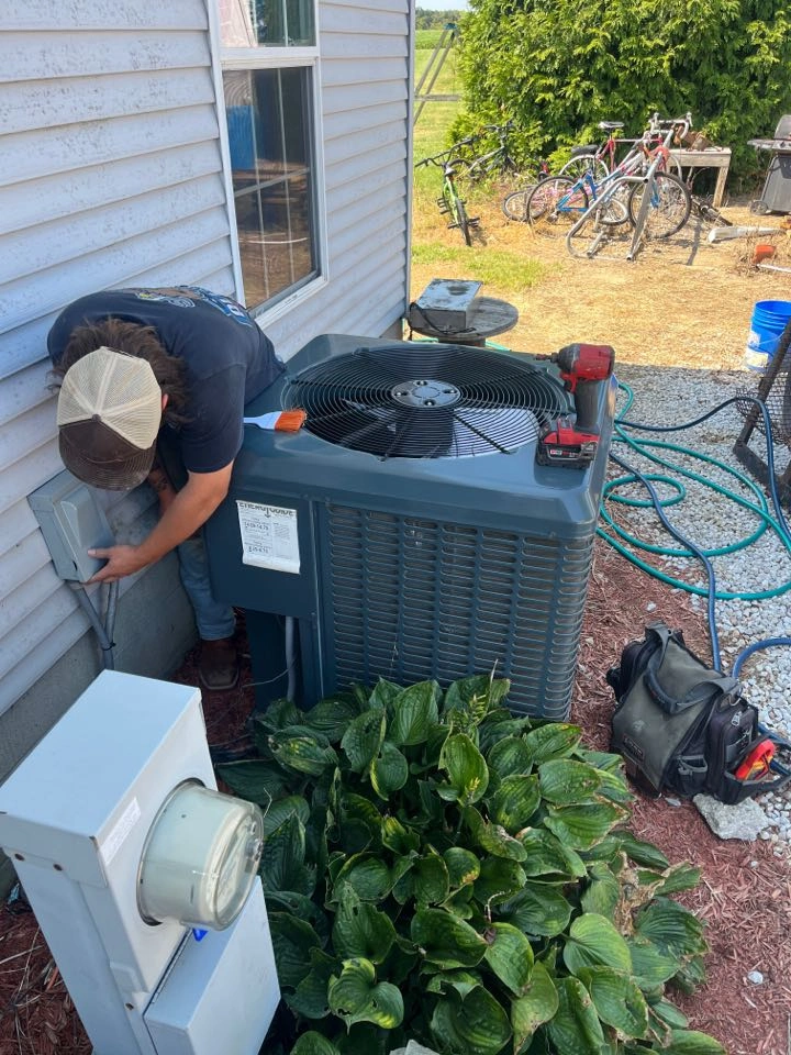 A person wearing a cap works on an outdoor air conditioning unit near a house.