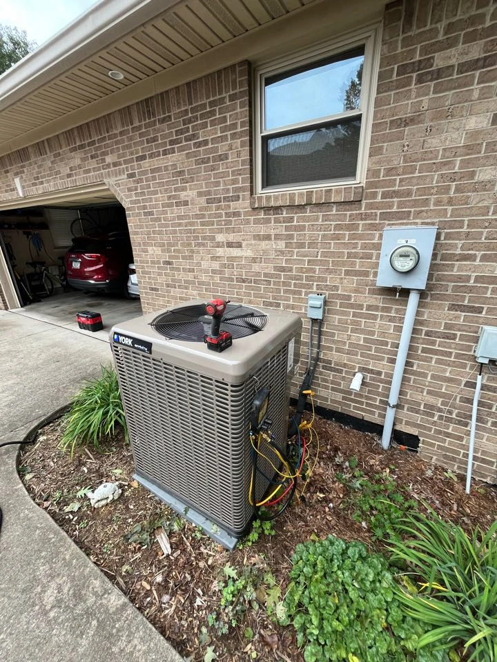Outdoor air conditioning unit with tools on top, set beside a brick house wall with a window and electric meter.