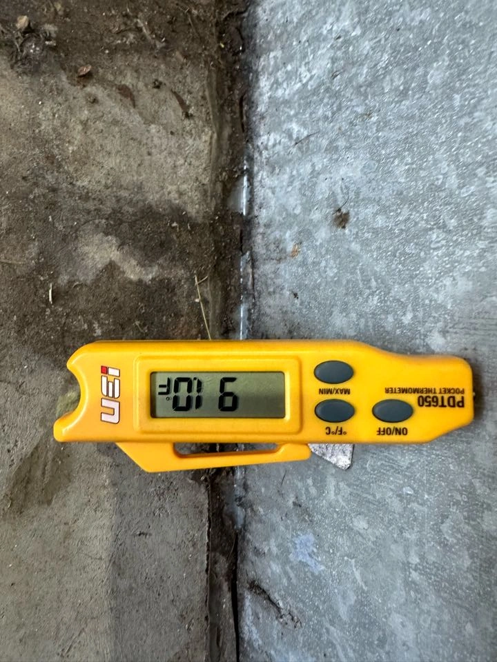 A yellow digital thermometer displays 91°F, resting on a concrete surface with soil beside it. 