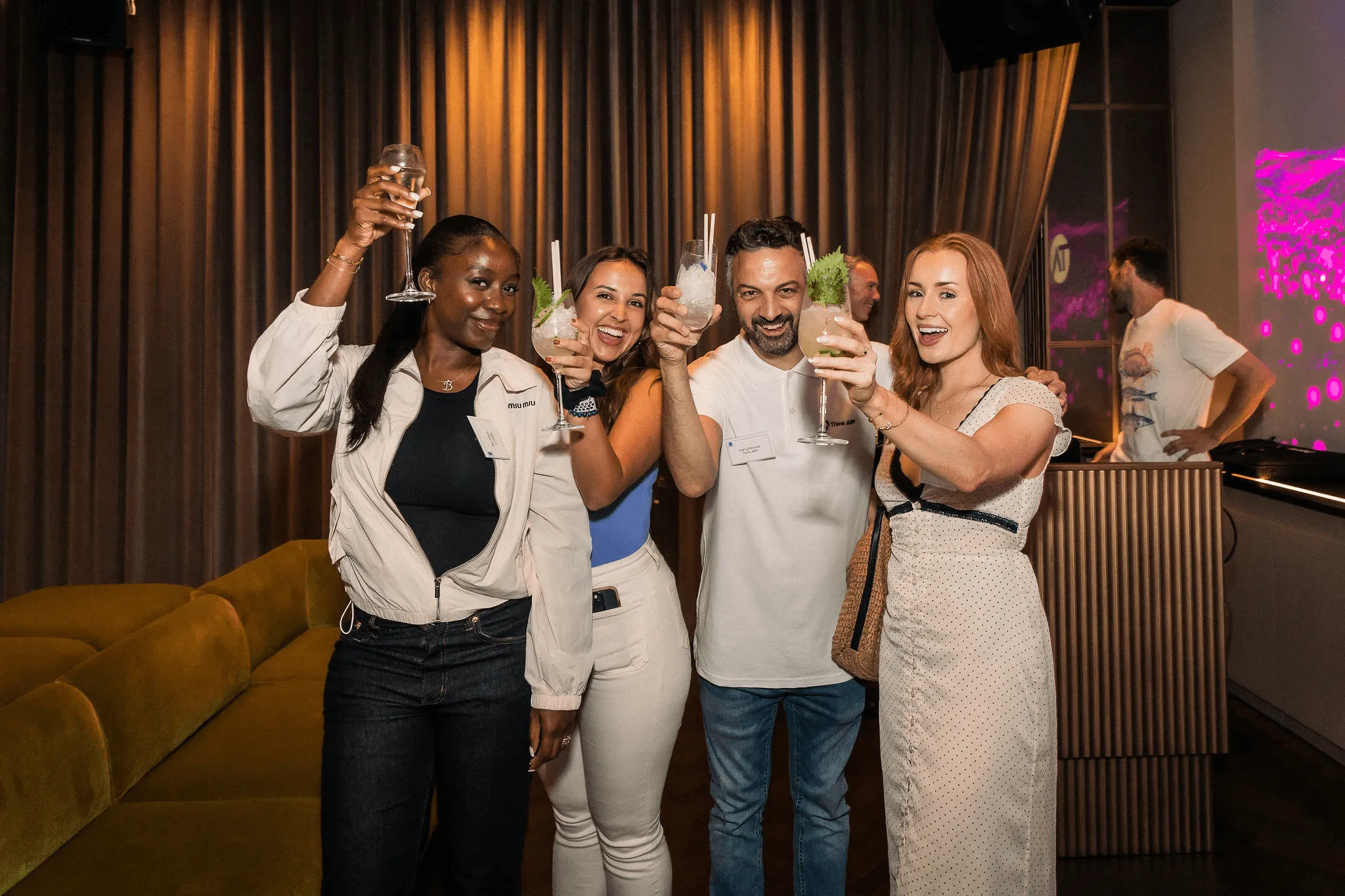 Four people smiling and raising drinks in a toast at an indoor event with dim lighting and curtains in the background.
