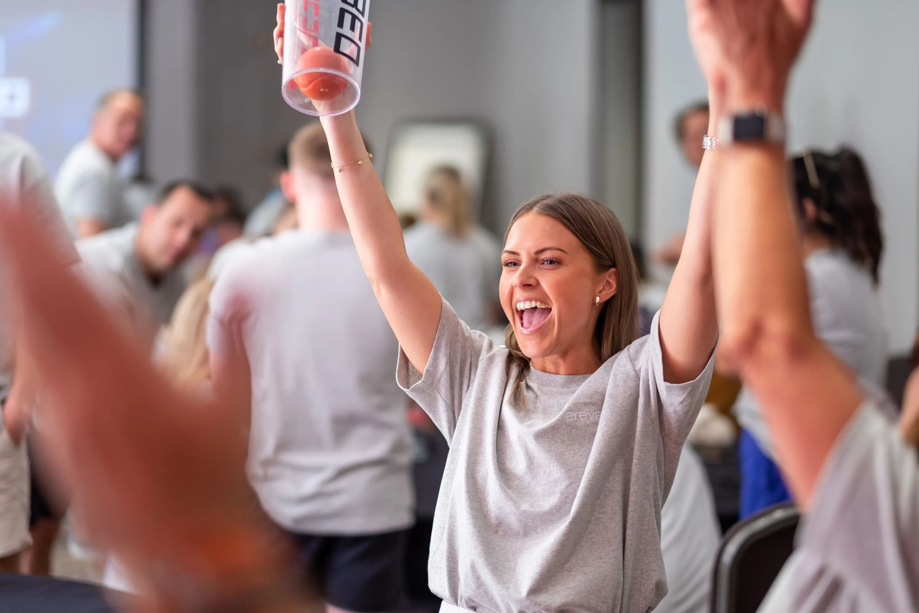 Woman cheering enthusiastically with arms raised, holding a clear shaker bottle with an orange ball inside, surrounded by people in a casual indoor setting.