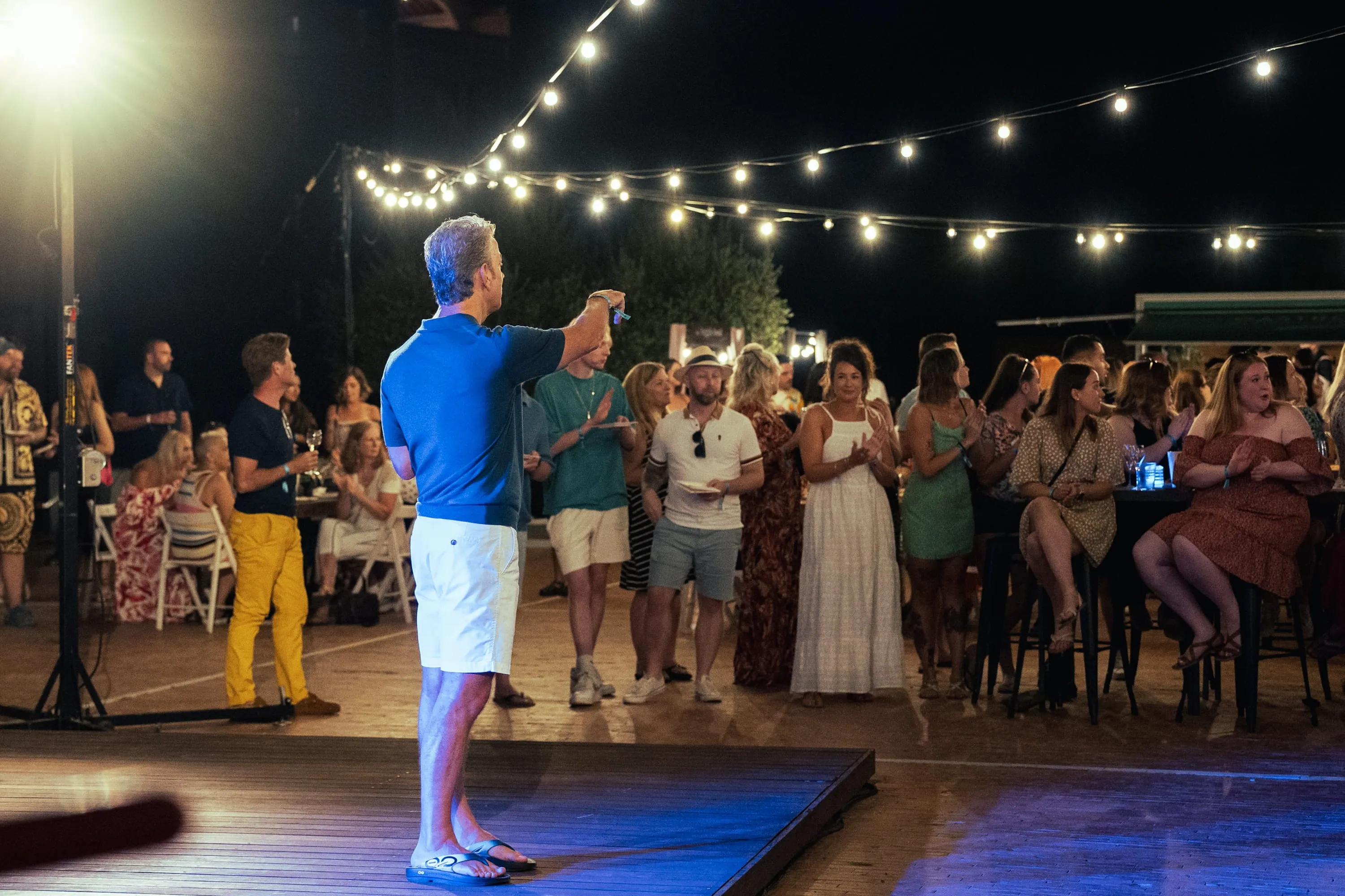 Man in a blue shirt and white shorts speaking to a crowd at an outdoor night event with string lights overhead.