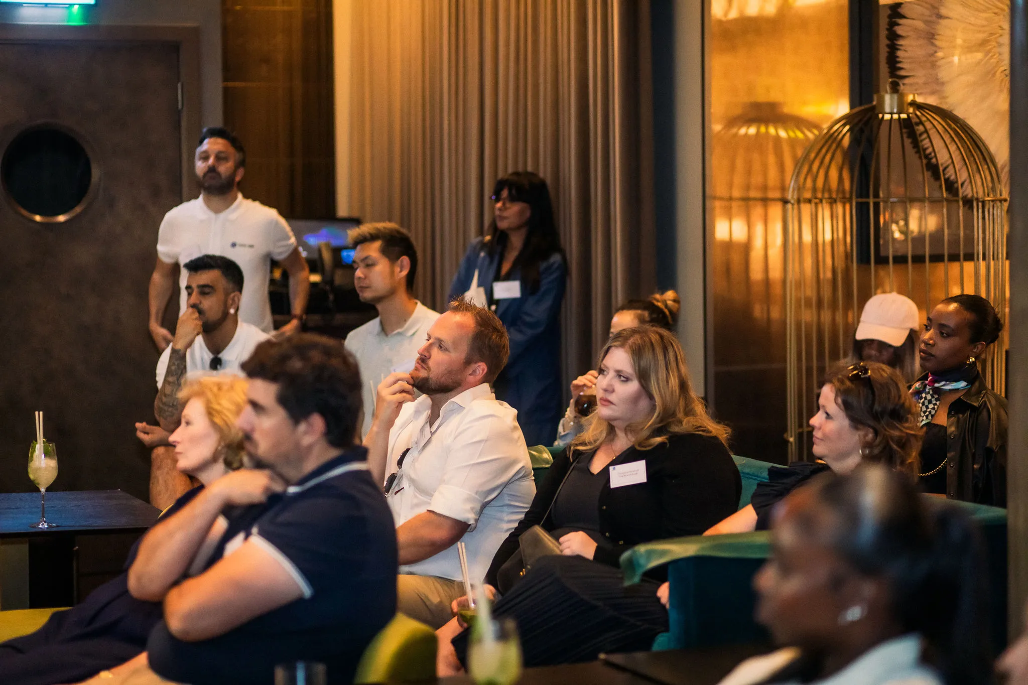 Group of diverse people seated and standing attentively listening in a dimly lit room with modern decor.