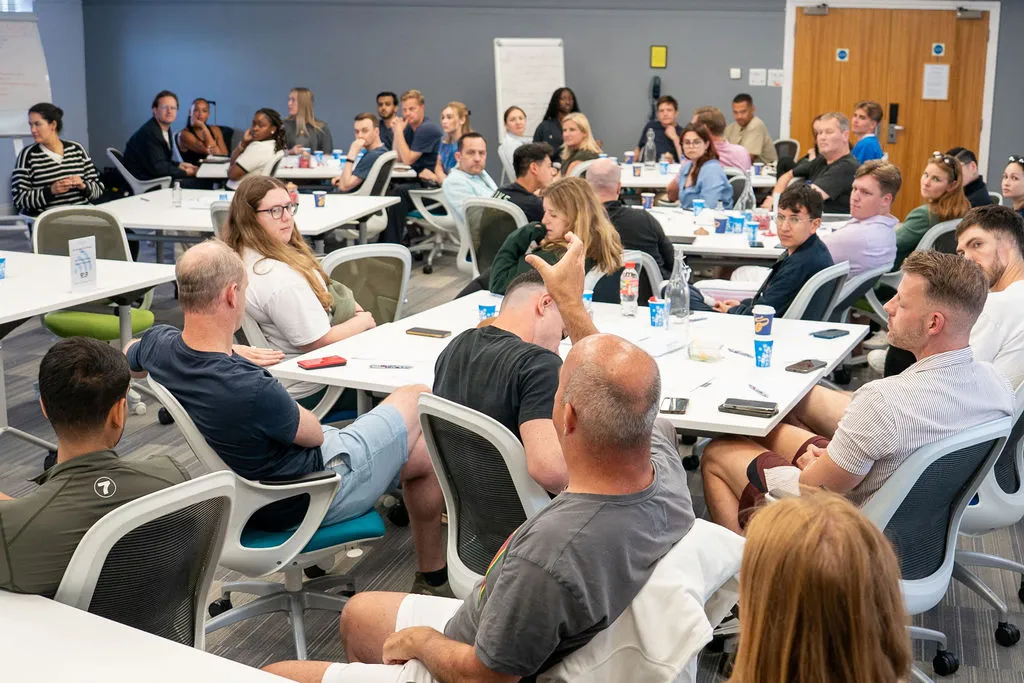 Group of adults seated around tables in a classroom setting engaged in a discussion.