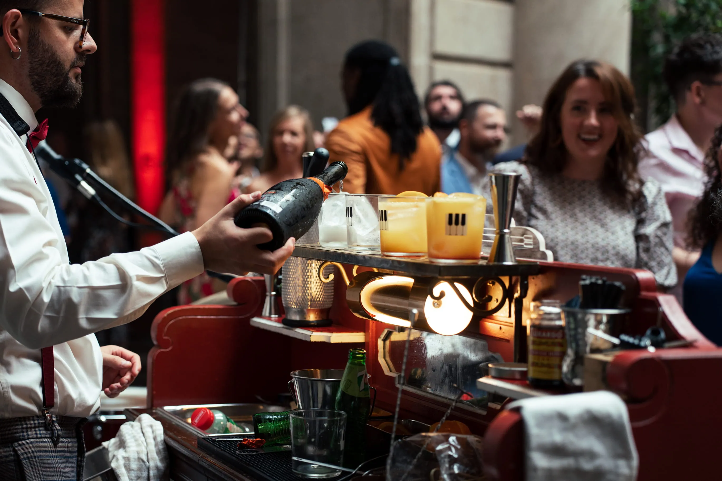 Bartender in white shirt and red bow tie holding a champagne bottle at a crowded event bar with mixed drinks and guests in the background.