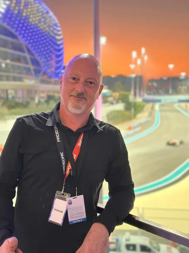 Man with a bald head and white beard wearing a black shirt and conference badges, standing near a railing with a racetrack and sunset sky in the background.
