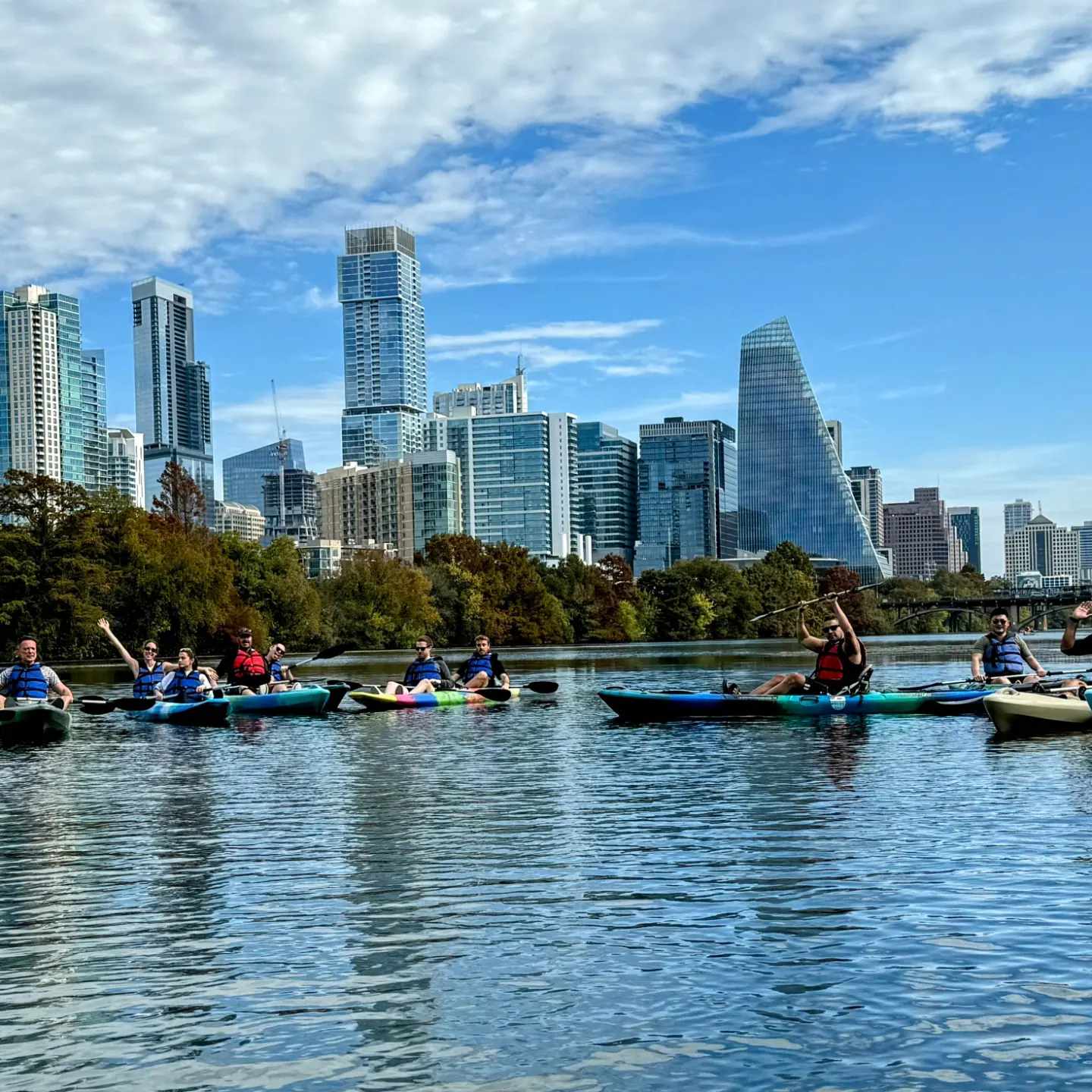 Group of people kayaking on a river with a modern city skyline and blue sky in the background.