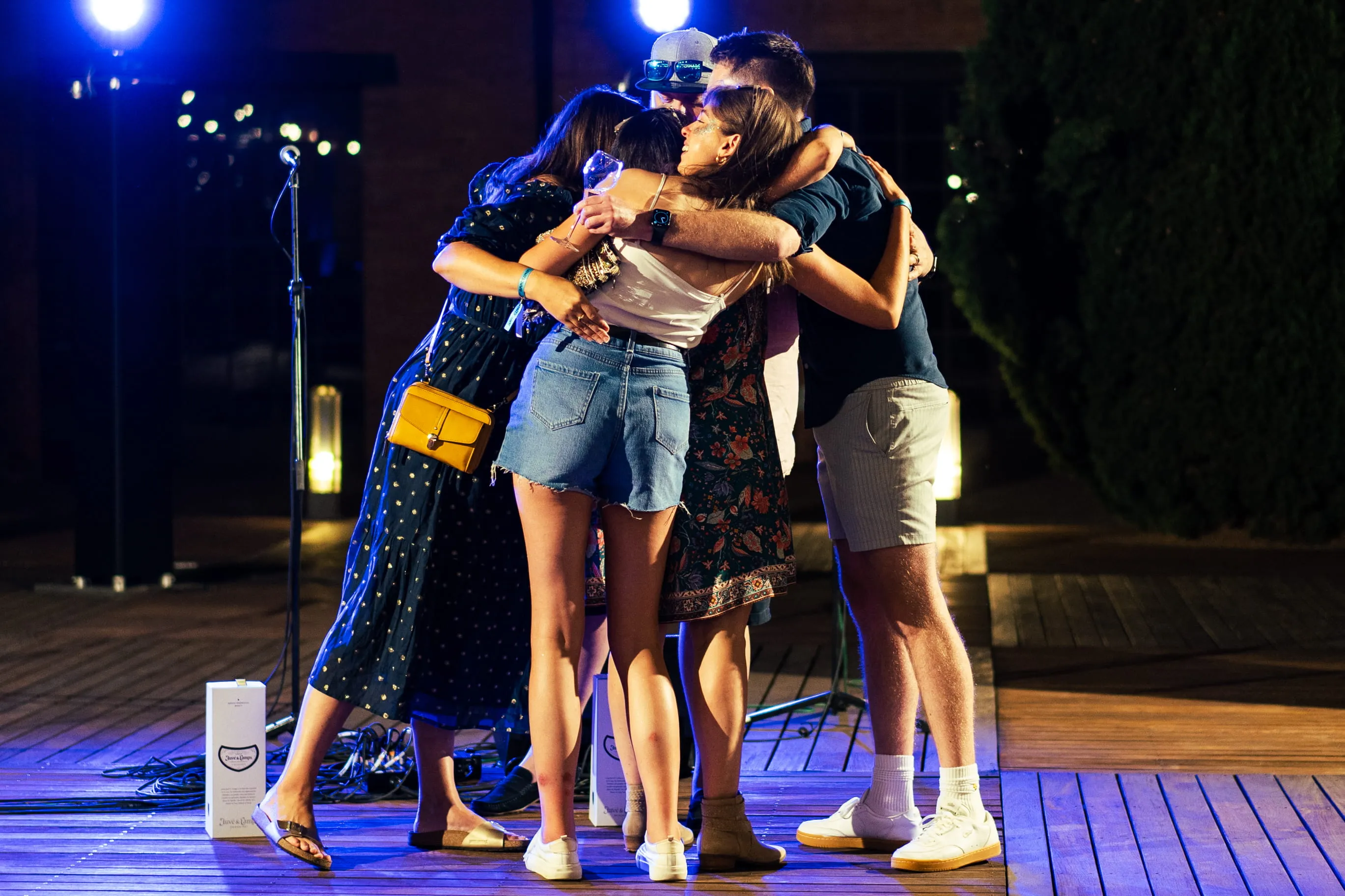 Group of five young adults embracing in a group hug during an outdoor nighttime event.