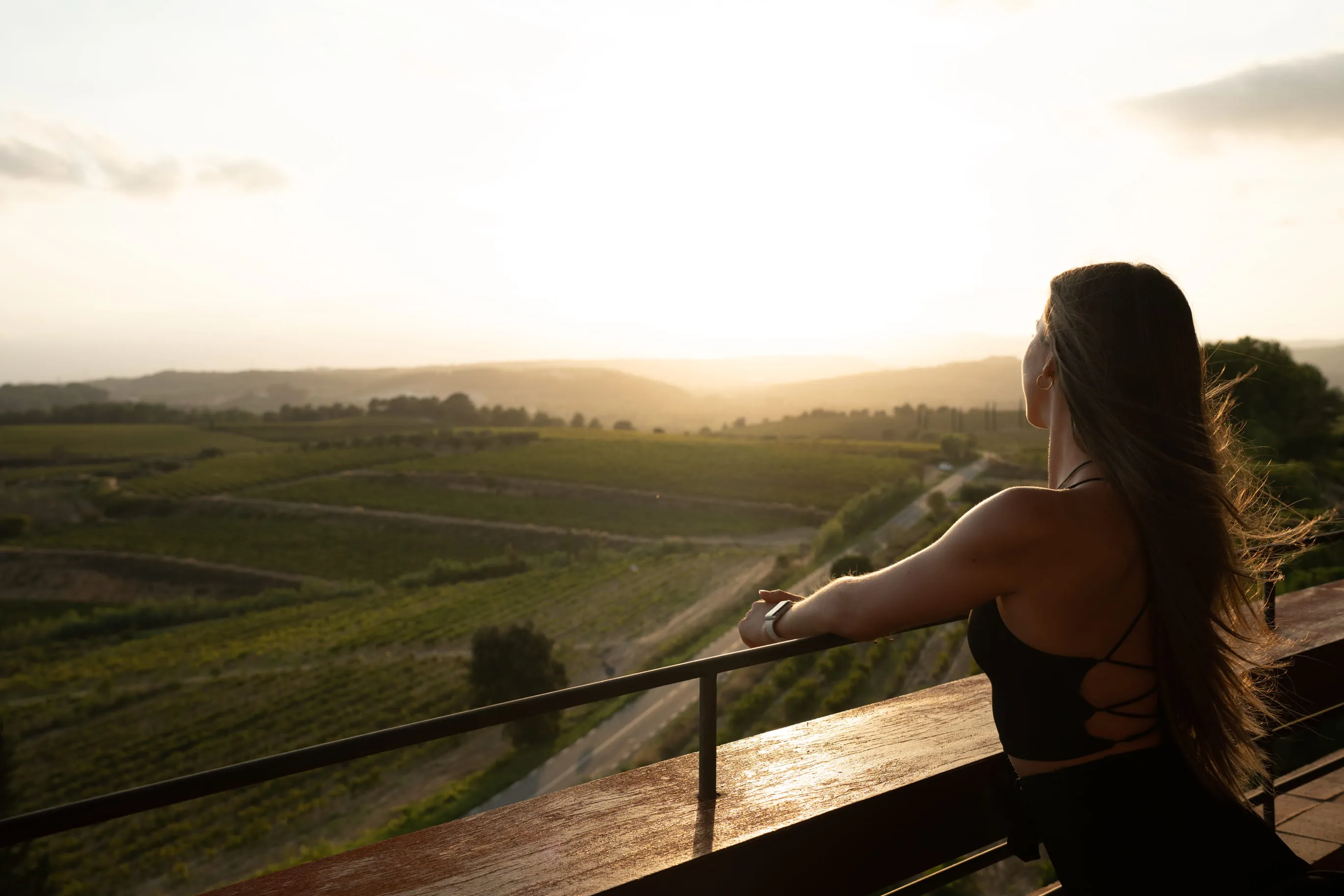Woman with long hair leaning on a wooden railing overlooking sunlit rolling vineyard hills at sunset.