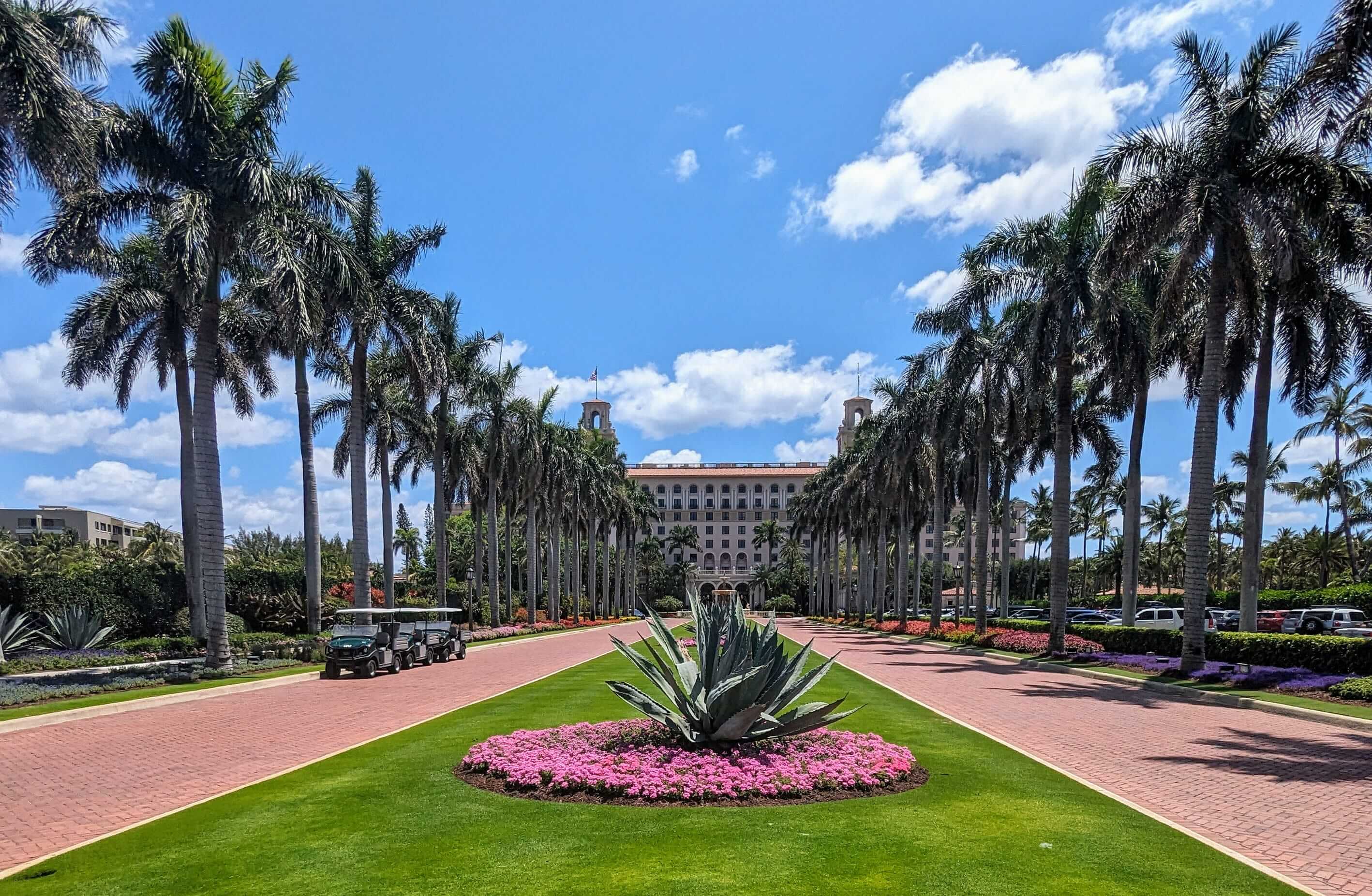 Wide brick driveway lined with tall palm trees leading to a large building under a blue sky with clouds.