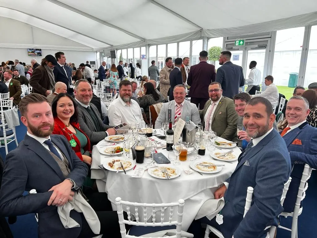 Group of men and one woman dressed in formal attire sitting around a round table with finished plates and drinks, inside a large event tent with other guests in the background.