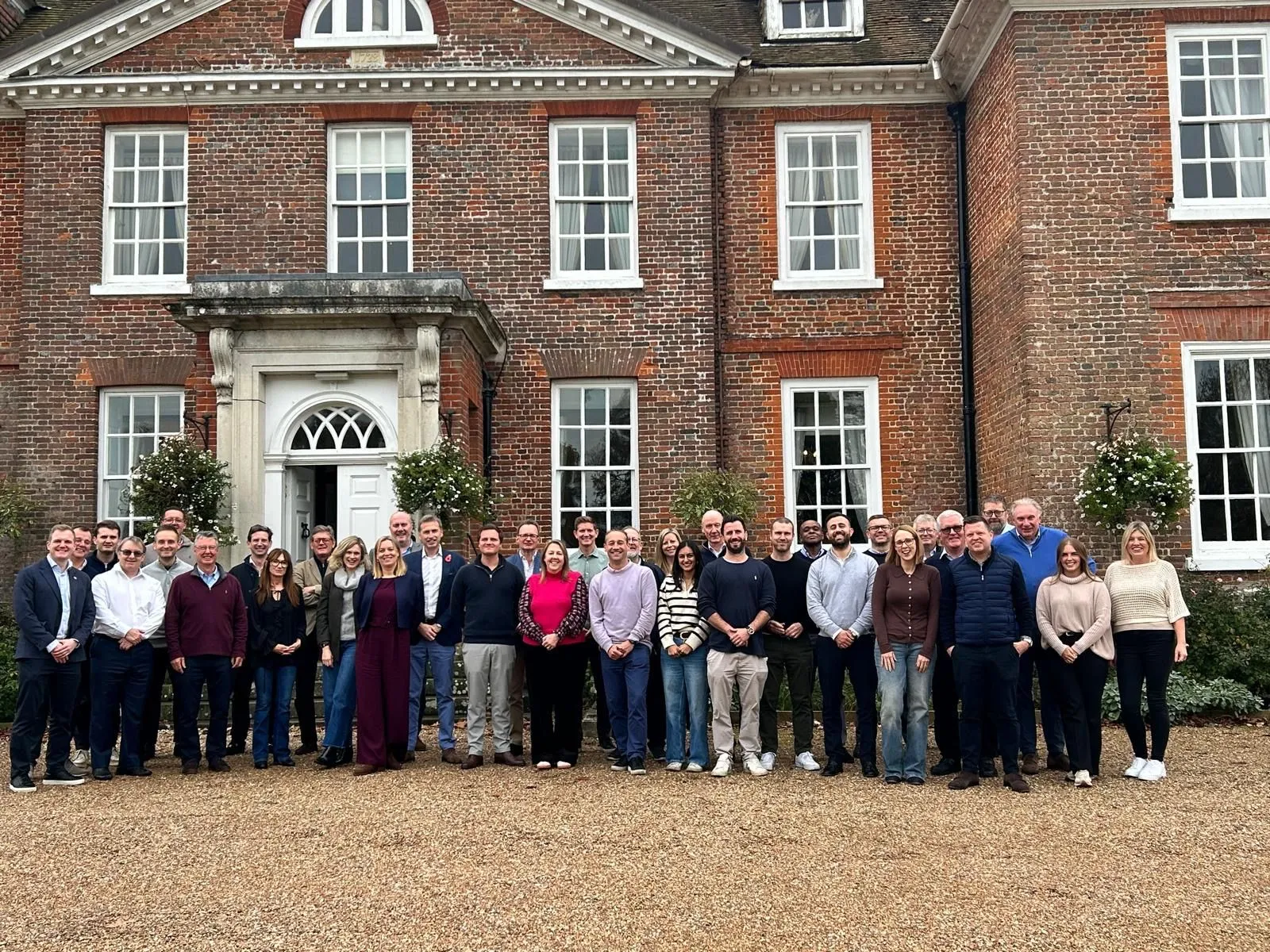 Large group of people posing in front of a historic brick building with white-framed windows and a central entrance.
