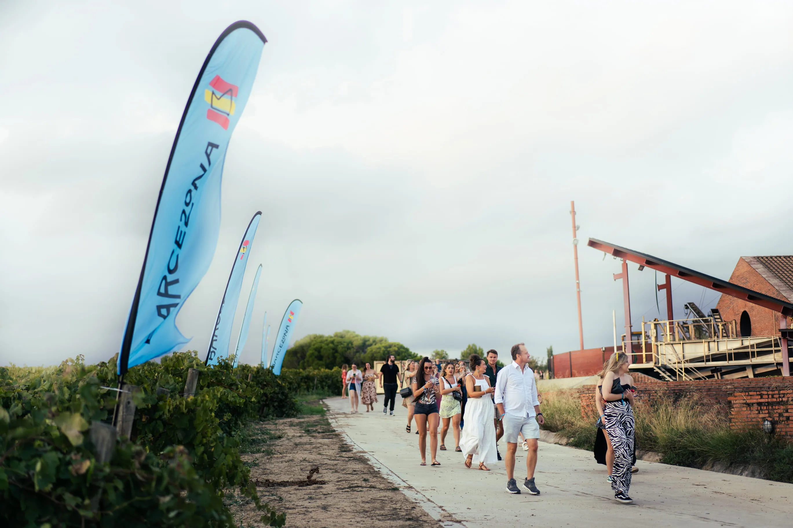 Group of people walking on a vineyard path with blue flags reading 'ARCEZONA' on the left and a brick building on the right.