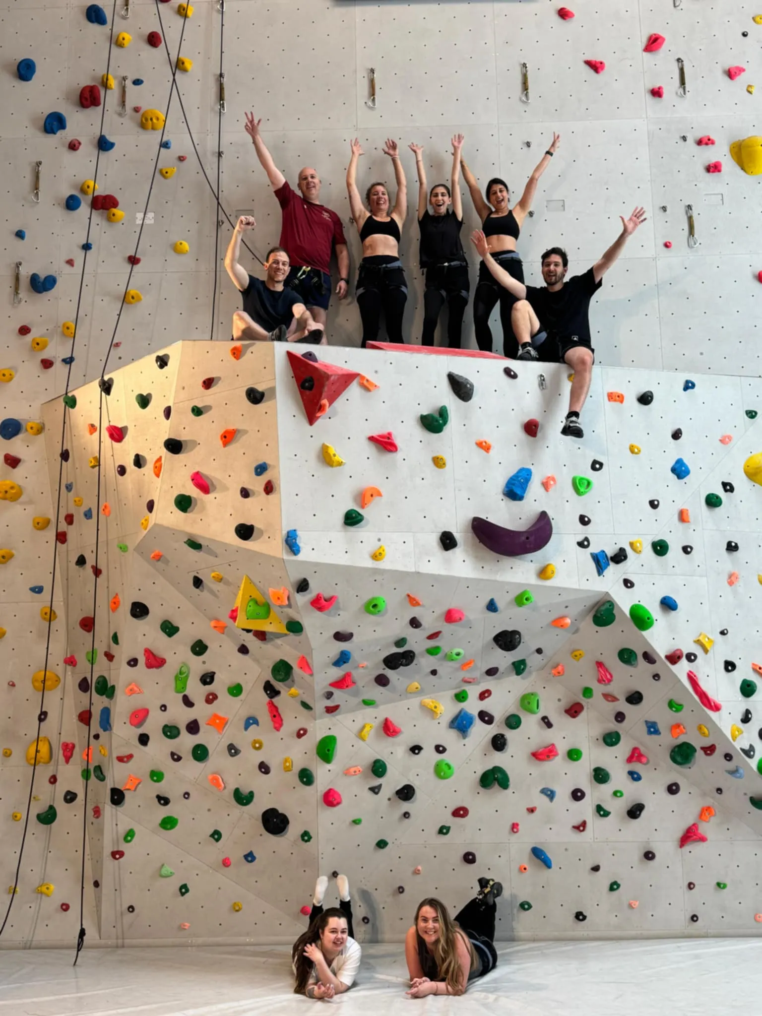 Group of nine people posing and smiling on and in front of a colorful indoor climbing wall.