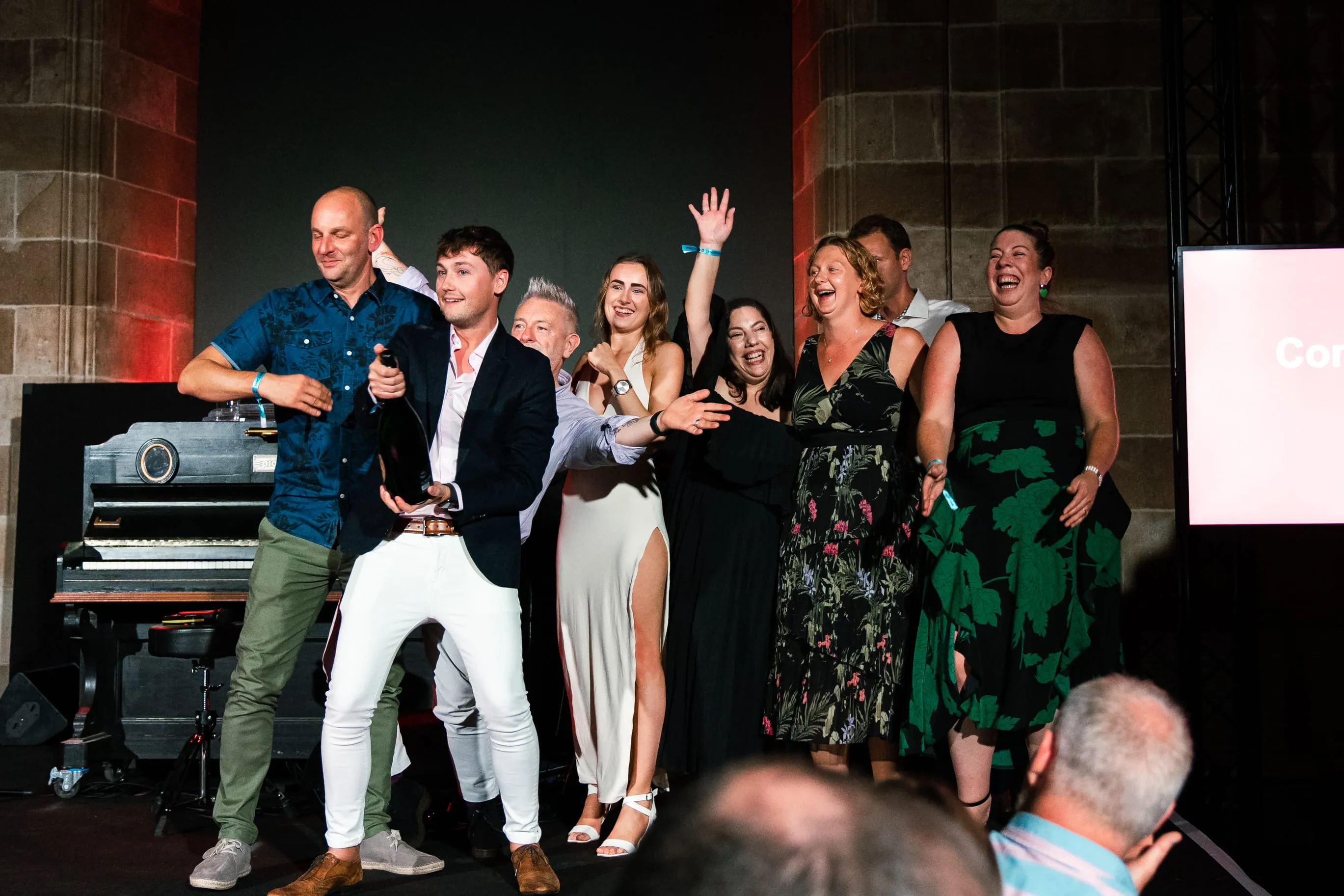 Group of people on stage celebrating with a man in front holding a bottle of champagne about to open it, everyone smiling and cheering.