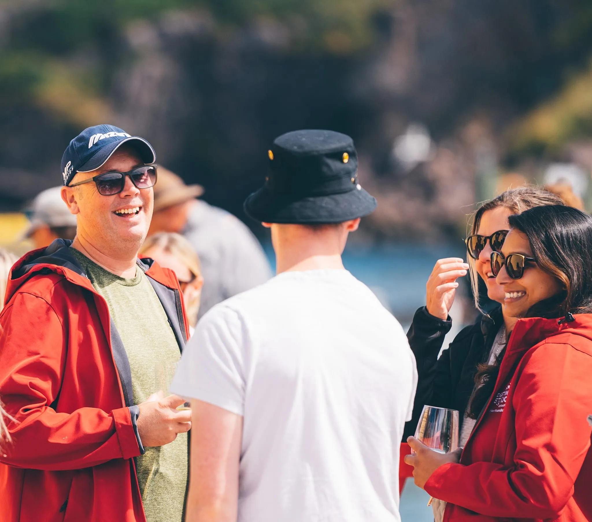 Group of people wearing sunglasses and jackets enjoying a sunny outdoor social gathering near water.