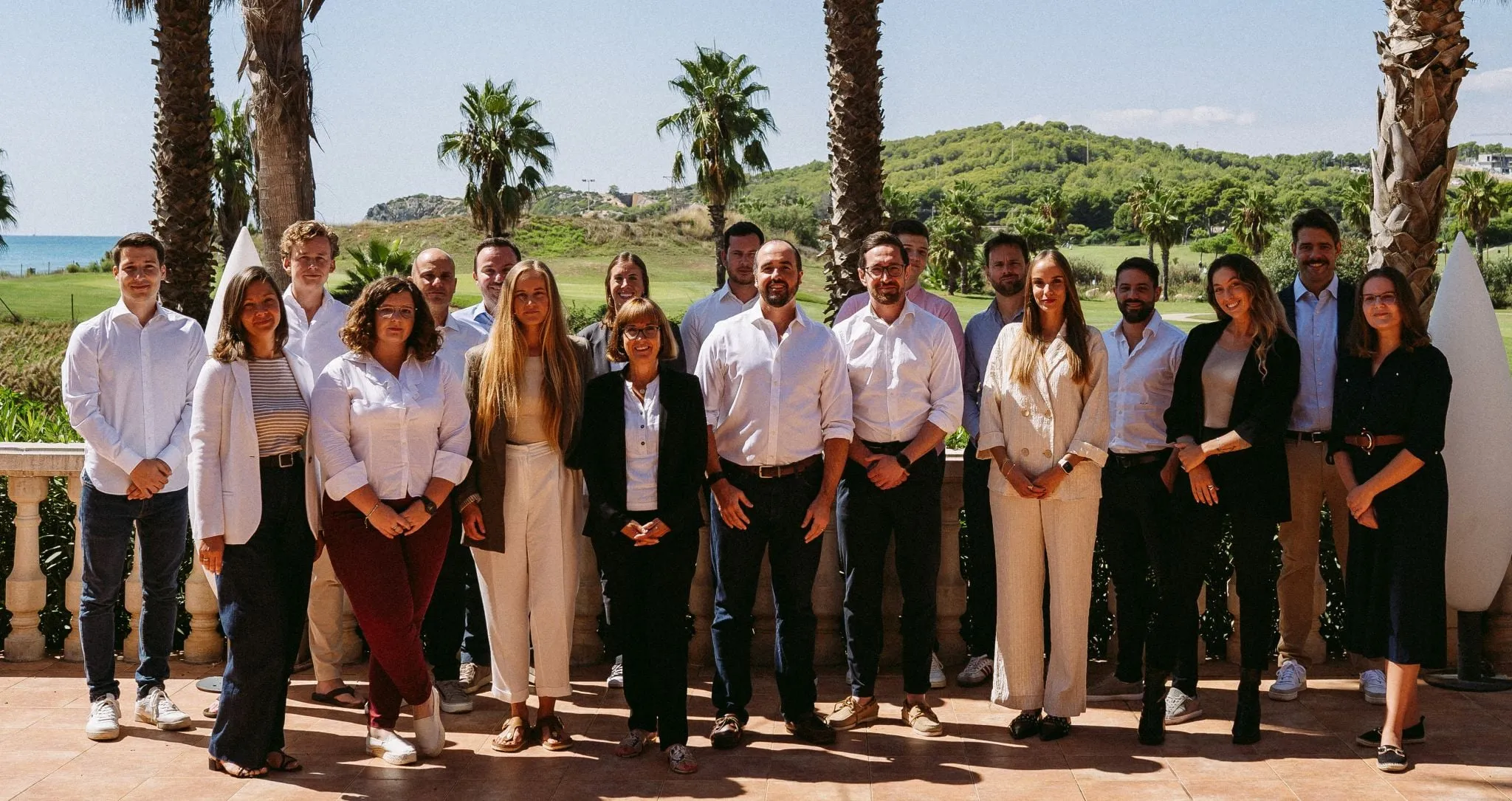 Group photo of twenty professionals standing outdoors on a terrace with palm trees and a green landscape in the background.