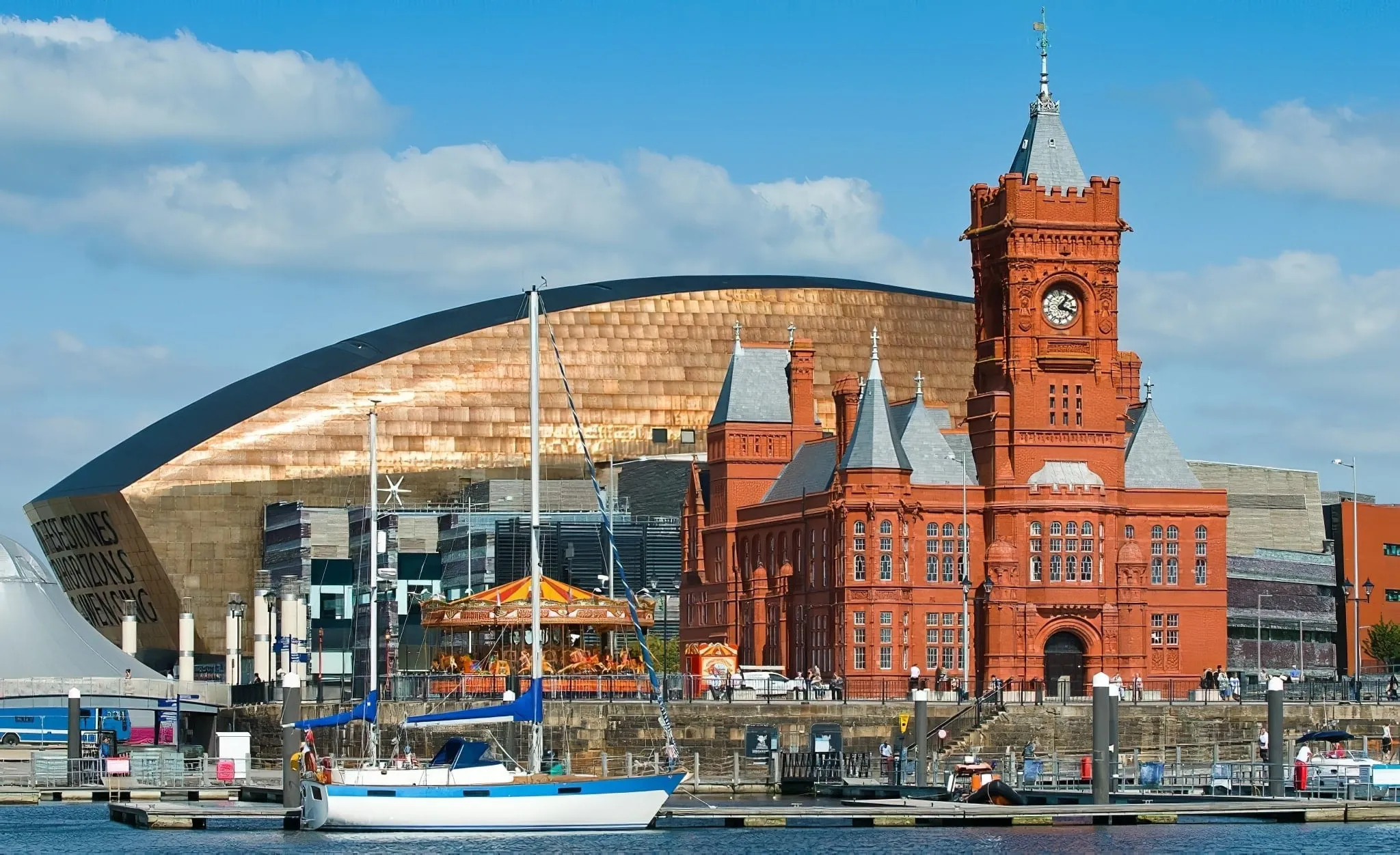 Waterfront scene in Cardiff featuring the red-brick Pierhead Building, a white sailboat in the water, and the modern golden Wales Millennium Centre in the background.