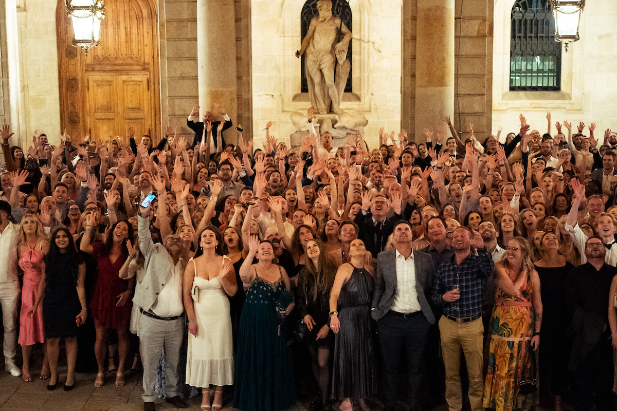 Large group of people dressed in formal attire raising their hands and looking upward inside a building with stone walls and statues.