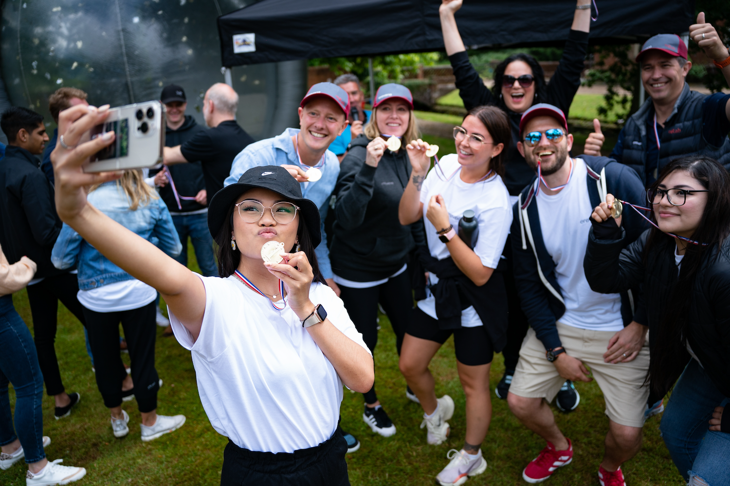Group of smiling people outdoors celebrating and showing medals while a woman in front takes a selfie.