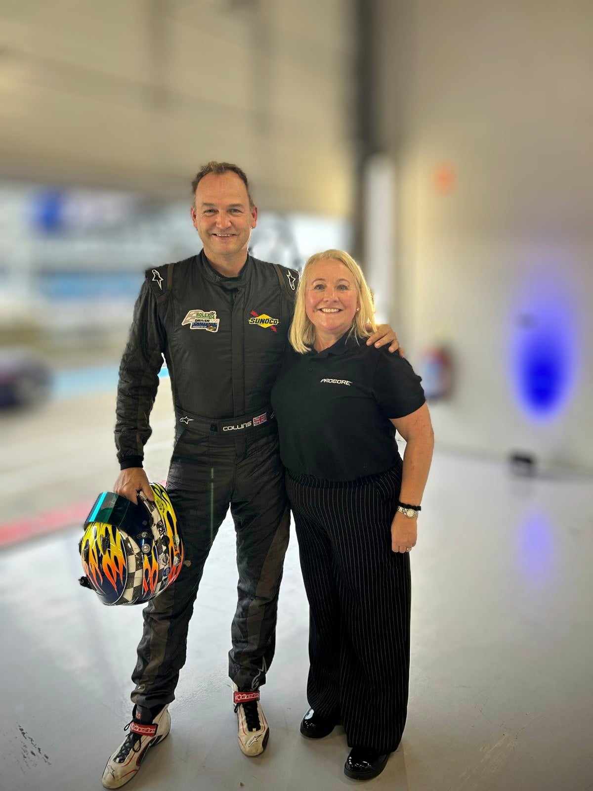 Man in black racing suit holding a colorful helmet stands next to a smiling woman in a black Procore shirt and pinstripe pants inside a garage.
