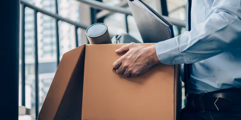 Person packt persönlichen Besitz in einen Umzugskarton, hält eine Kaffeetasse und Unterlagen.