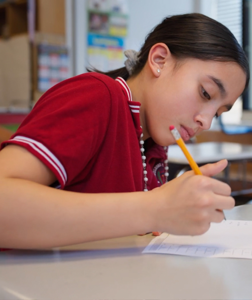 Student in a red shirt writing with a pencil on paper at a classroom desk.