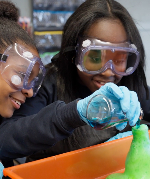 Two girls wearing safety goggles and gloves pouring blue liquid into a green foam experiment.