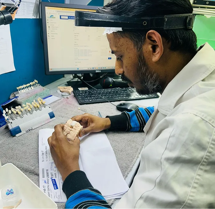 Dental technician examining a dental mold at a workstation with a computer and shade guide tools.