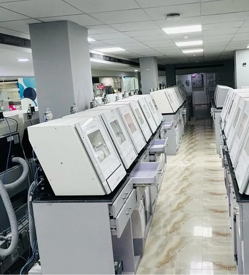 A laboratory with two rows of white automated analyzers facing each other along a tiled floor corridor.