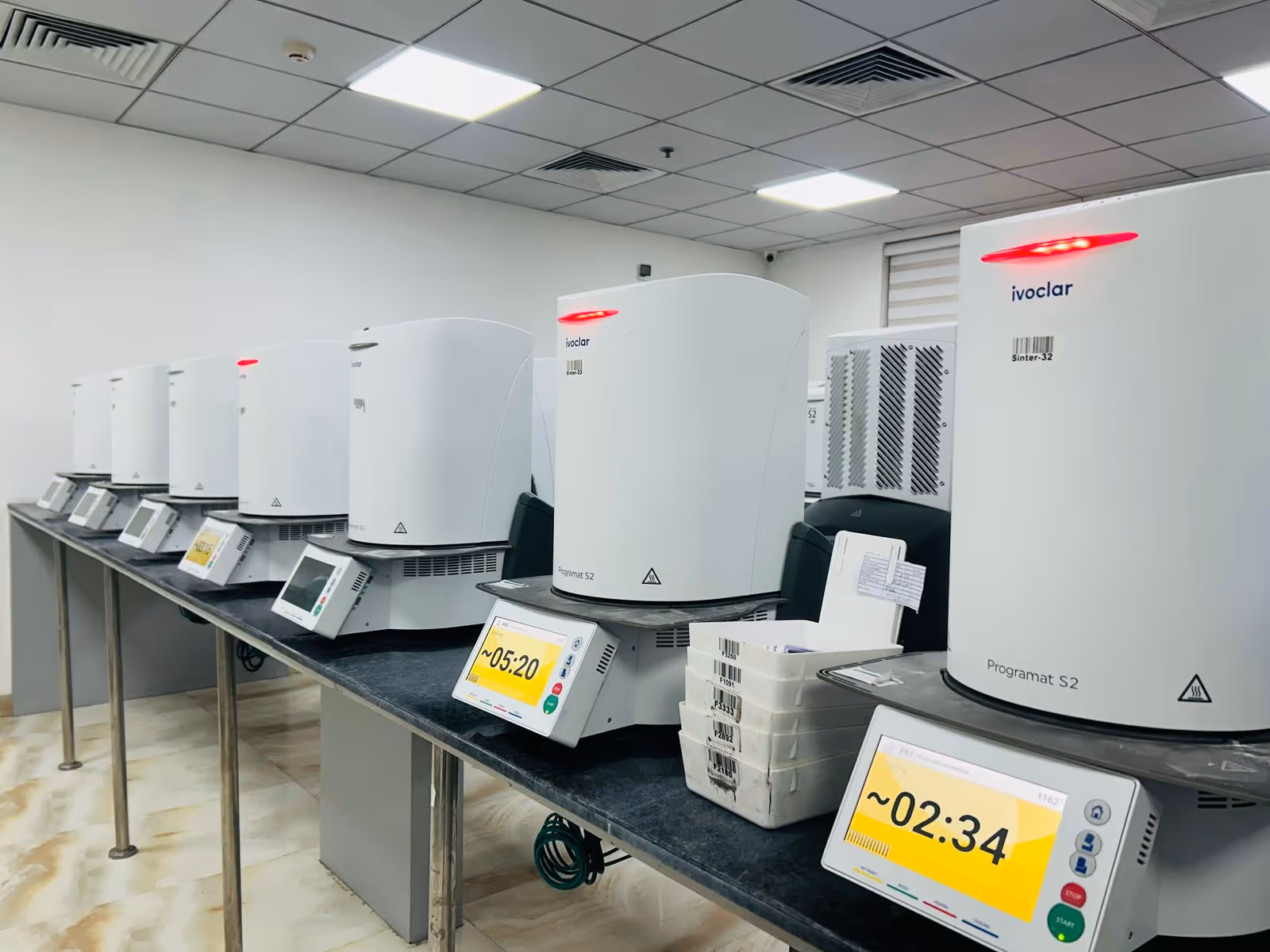 Row of white Ivoclar Programat S2 dental ceramic furnaces with digital timers on a long black counter in a lab.