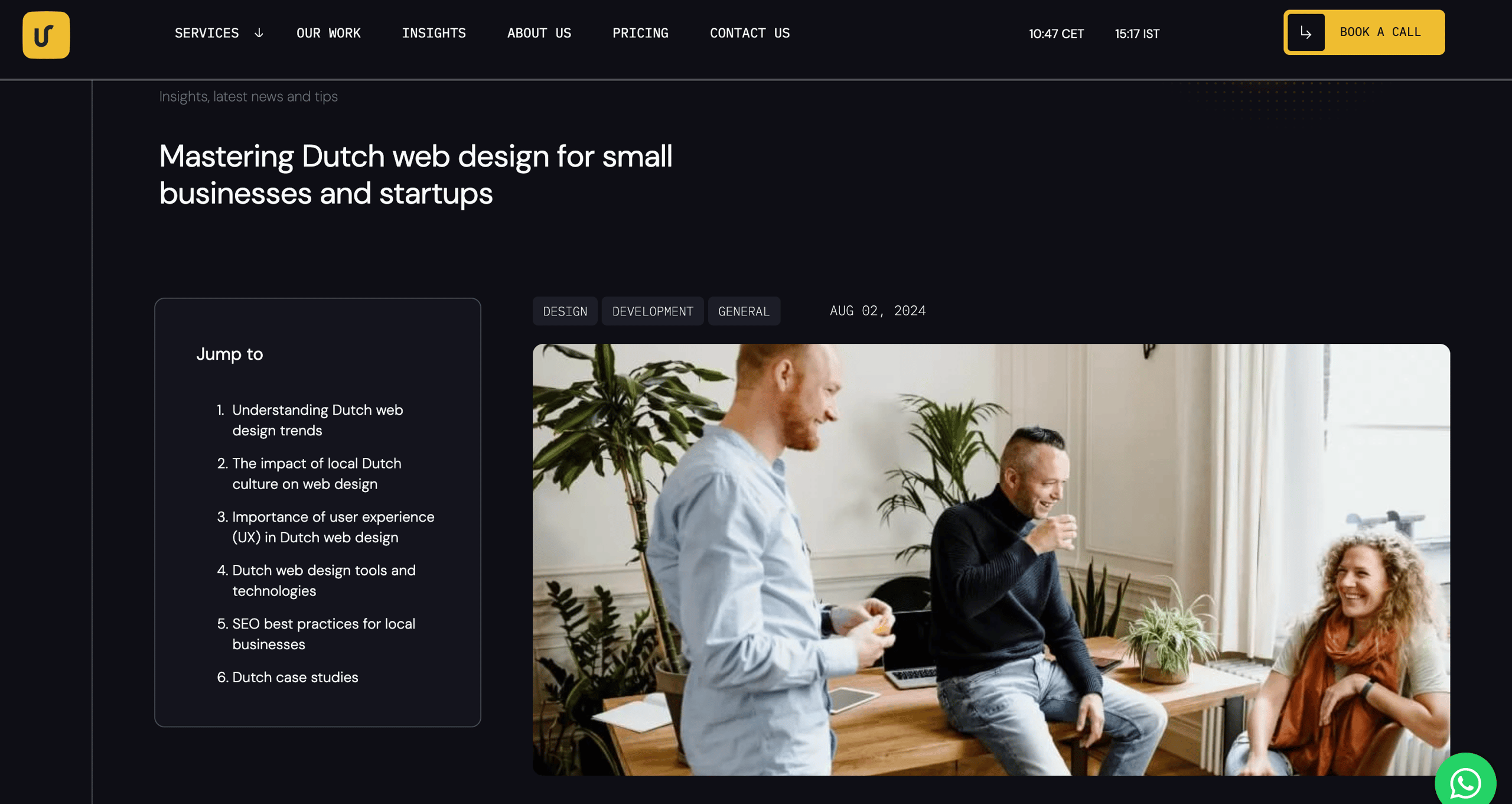 Three people casually chatting and laughing around a wooden table in a bright office with plants.