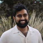 Man with black curly hair and beard smiling outdoors with tall grass in the background.