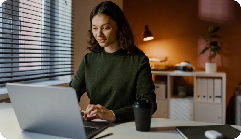 Young woman in a dark green sweater working on a laptop at a desk with a coffee cup nearby, a warmly lit office background with shelves and a plant.